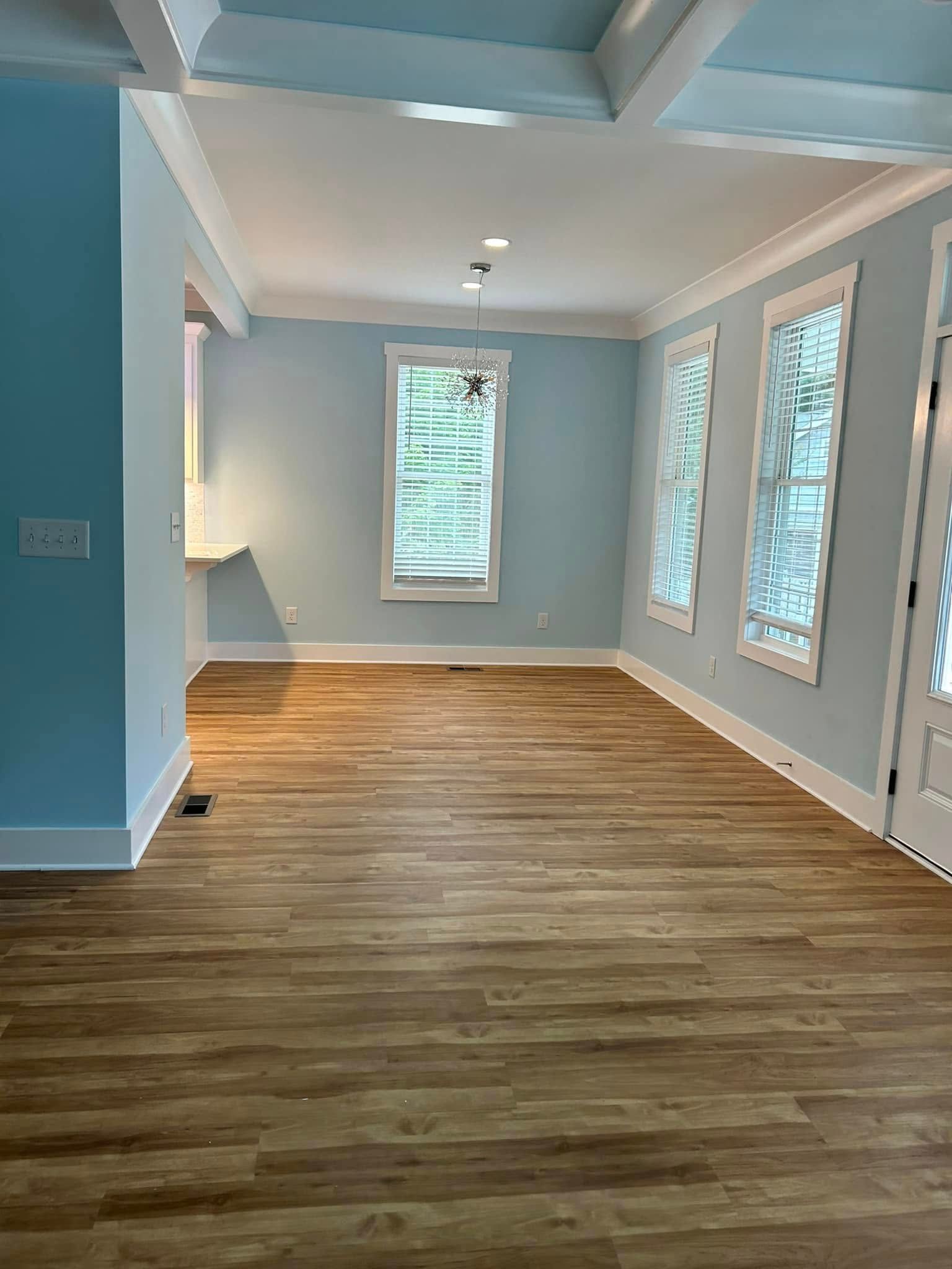 Empty room with light blue walls, white trim, and wood-look flooring. Natural light streams in windows.