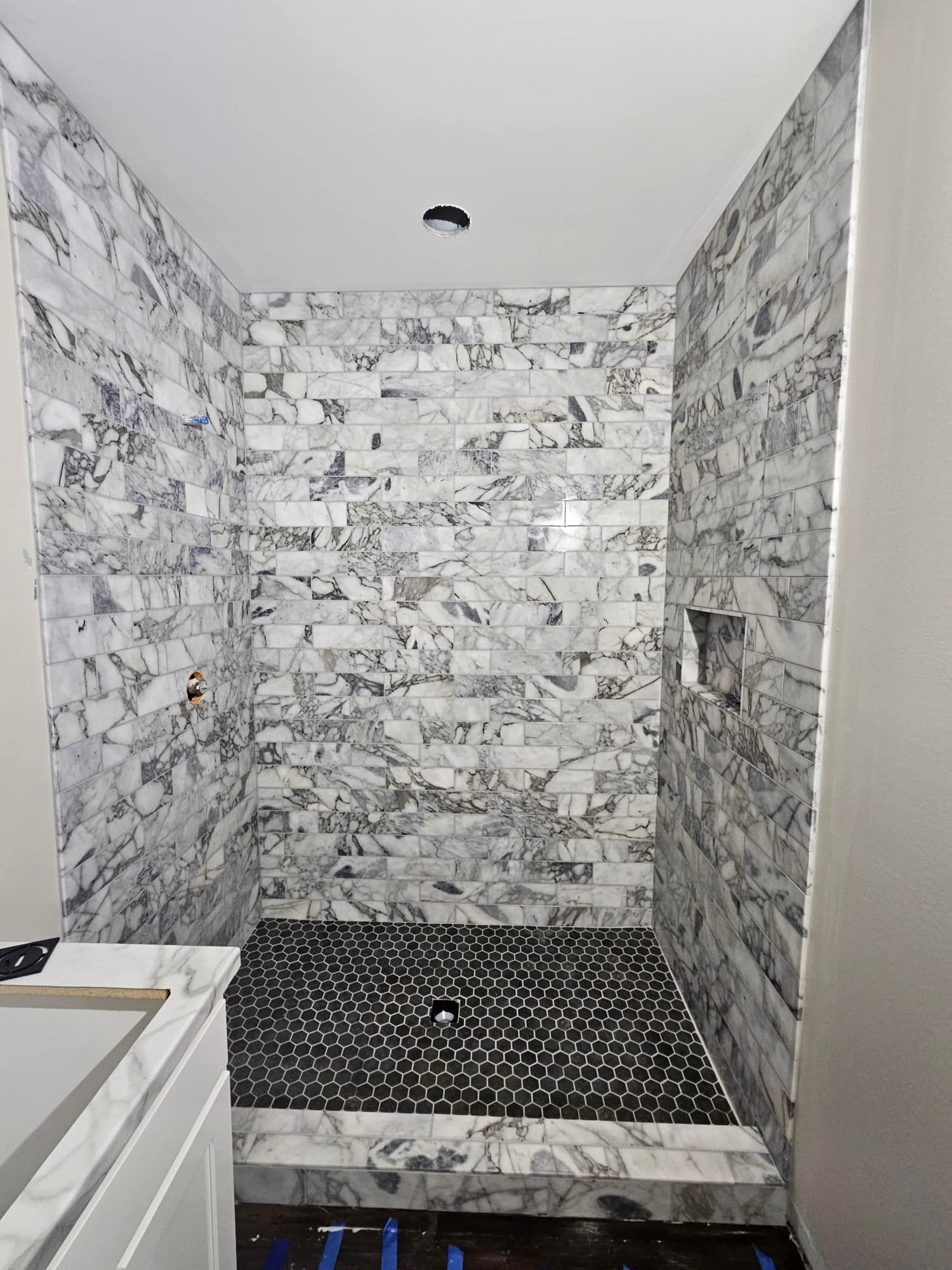 Shower with marble-patterned tile walls and black pebble tile floor. Recessed shelf on right wall.
