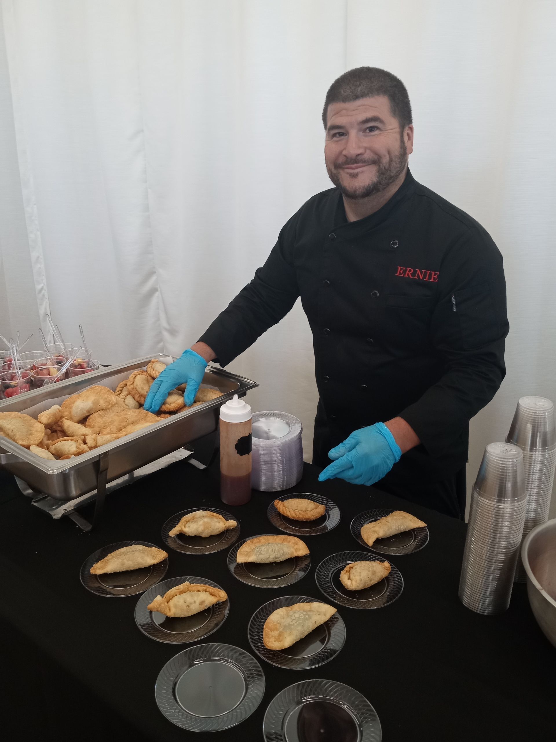 A man wearing blue gloves is standing in front of a table full of food.