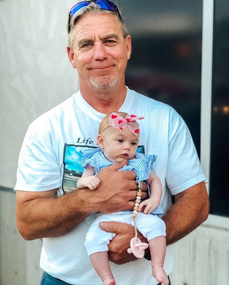 Man in white shirt holding a baby with a heart bow. They are outside. The man is smiling.