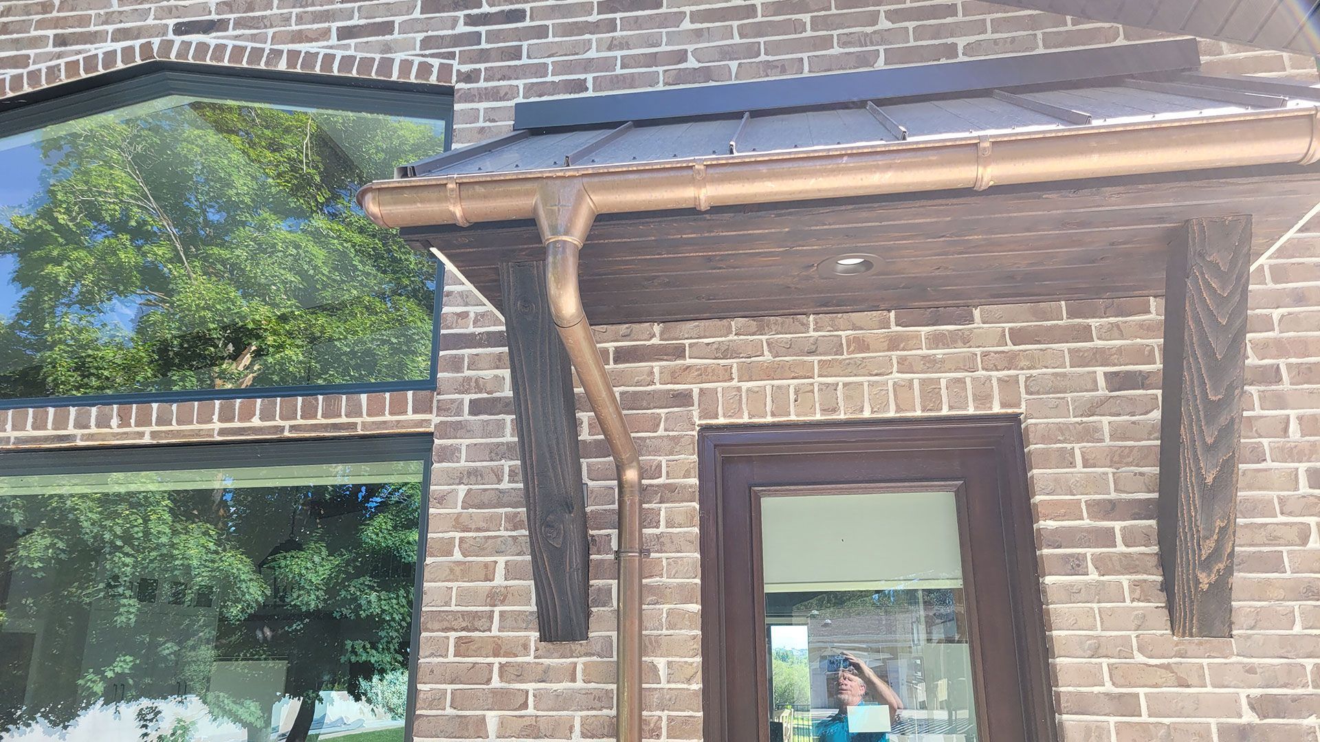 Brick building with copper gutters, a dark wooden canopy over a door, and large windows reflecting trees.