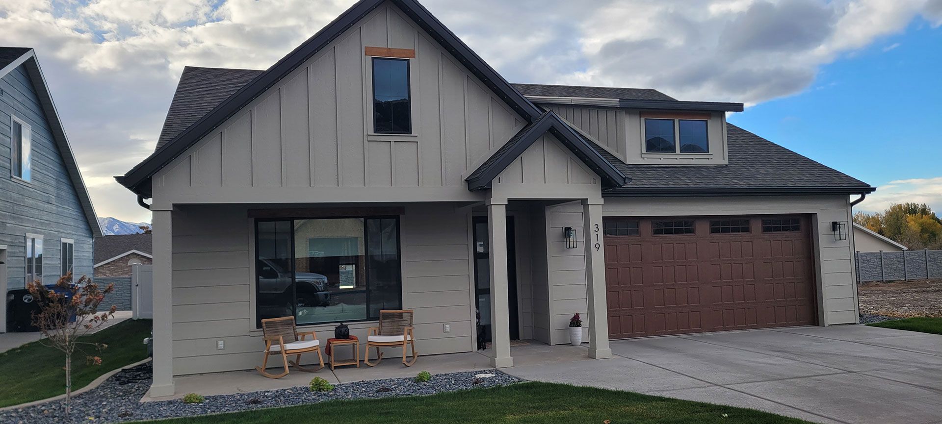 A light gray house with a brown garage door. Two chairs sit on the porch.