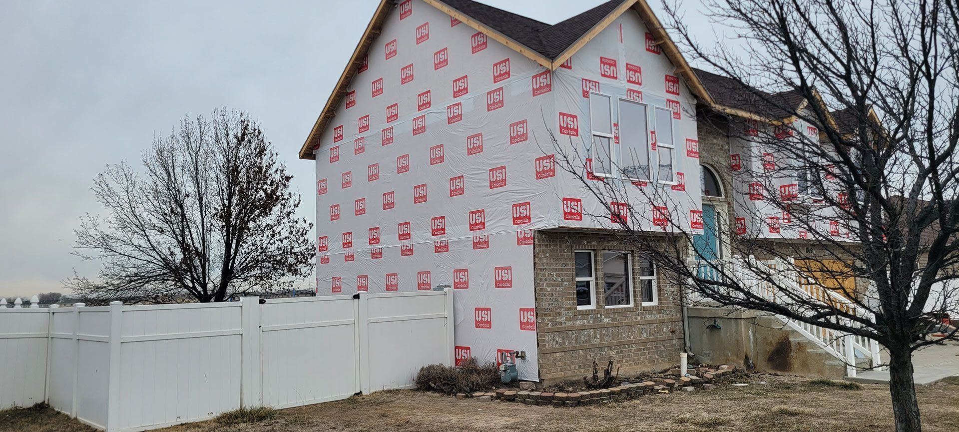House under construction with white siding and stone veneer, surrounded by a white fence and barren trees.