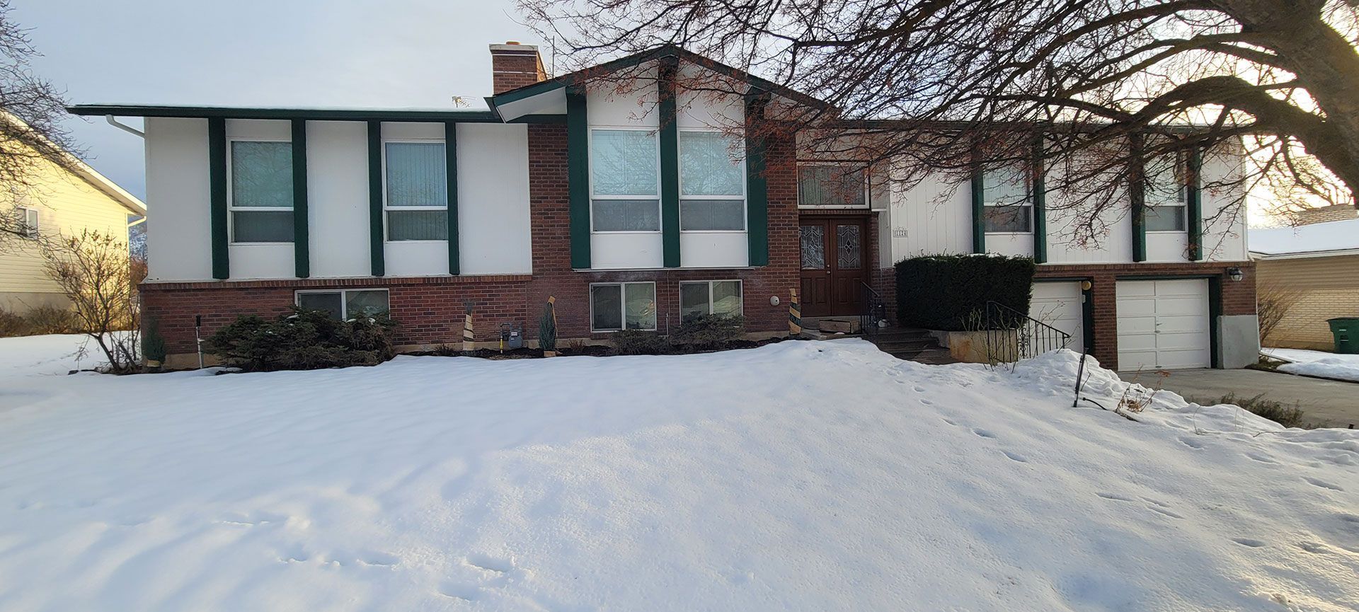 A brick and white split-level house covered in snow with a bare tree in front.