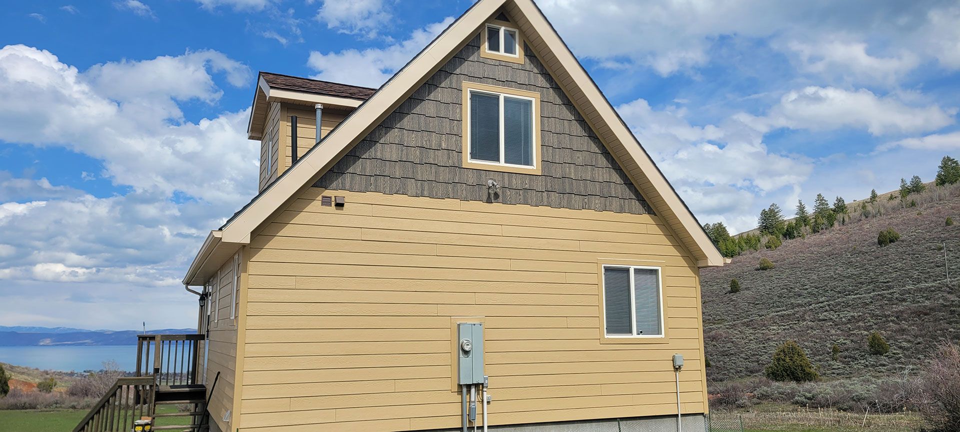 A two-story house with tan siding and a dark gray gable, set against a blue sky with clouds.