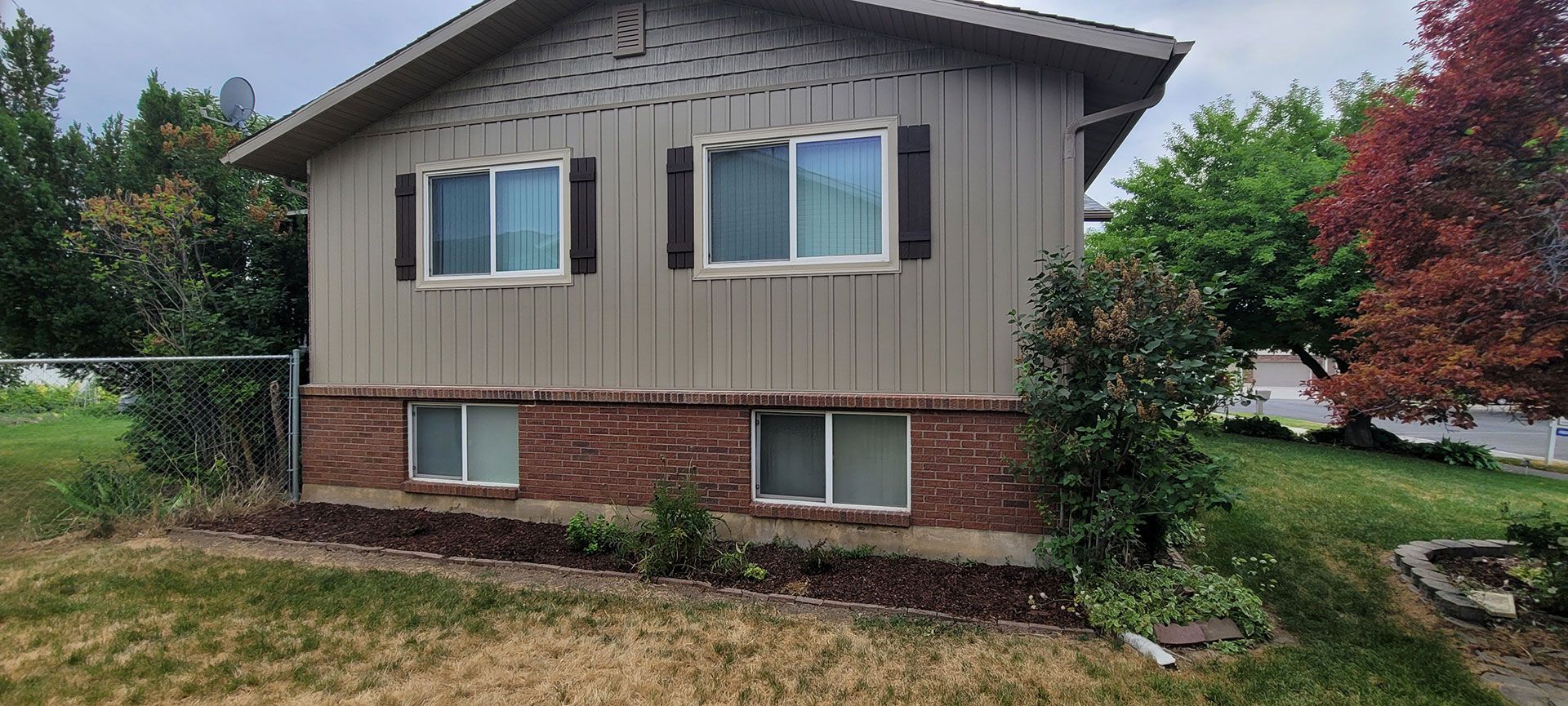 Tan house with brick foundation, windows, and dark shutters. Green yard with trees. Overcast sky.