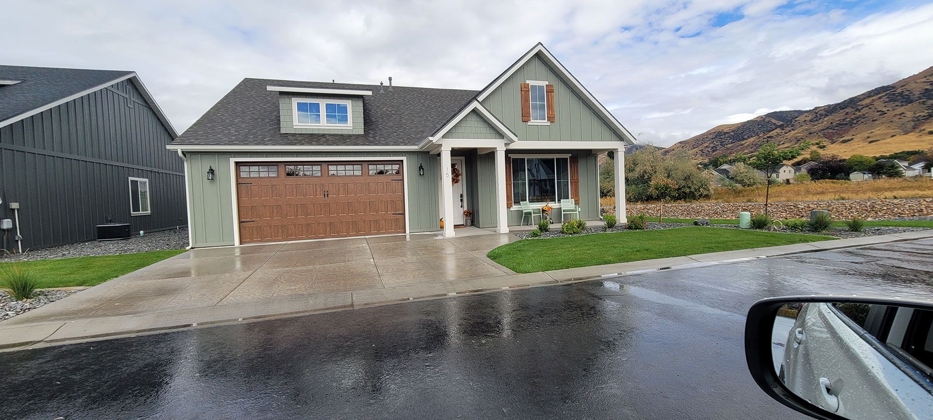 House with green siding and brown garage door on a wet, paved road; mountains in the background.
