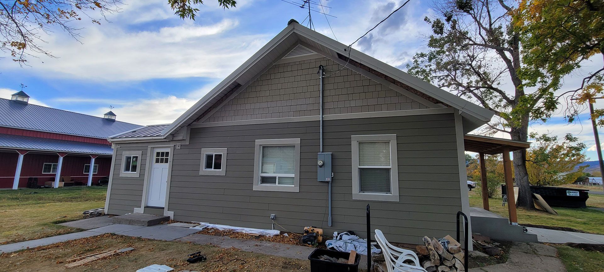 Gray house with white door and windows, siding, and porch. Red barn in the background under a blue sky.