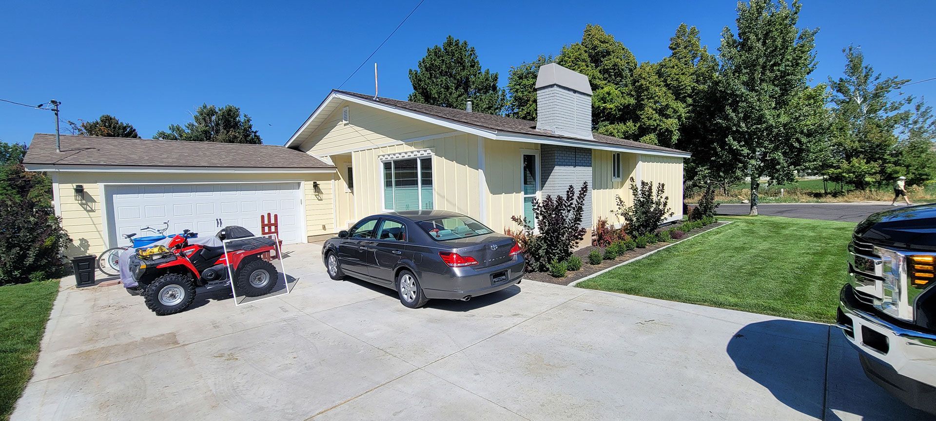 Yellow house with a gray car, a red ATV, and a black truck parked in the driveway on a sunny day.
