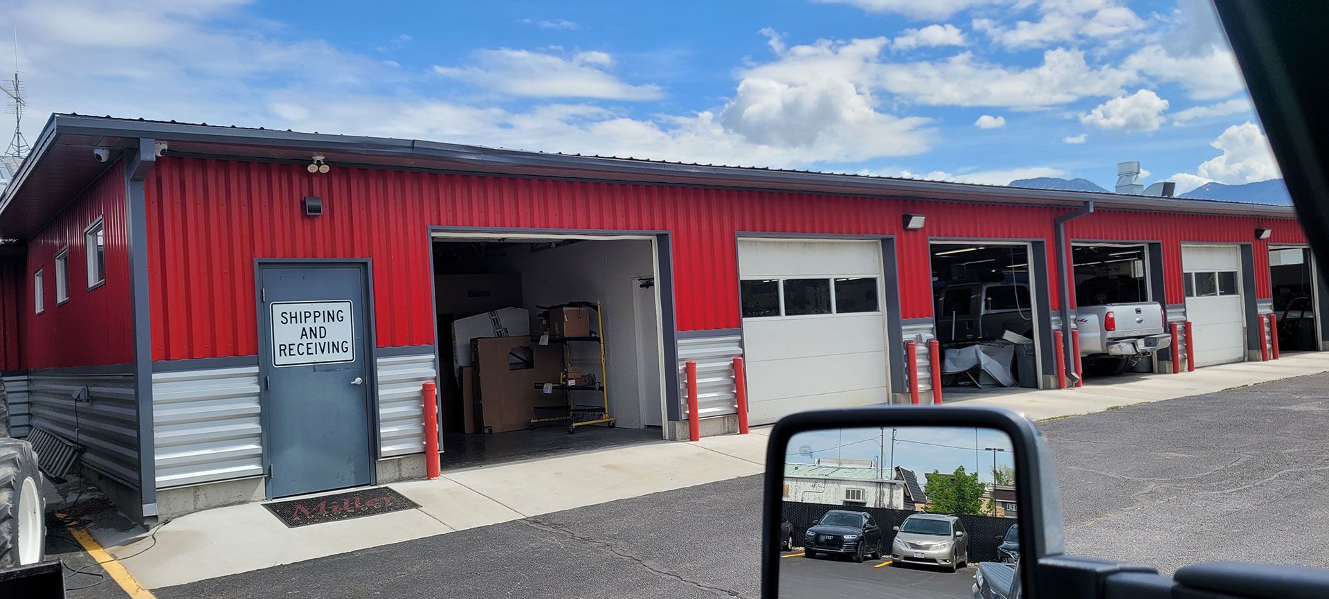 Exterior view of a red automotive garage with open bays, parked vehicles, and a reflective side mirror.
