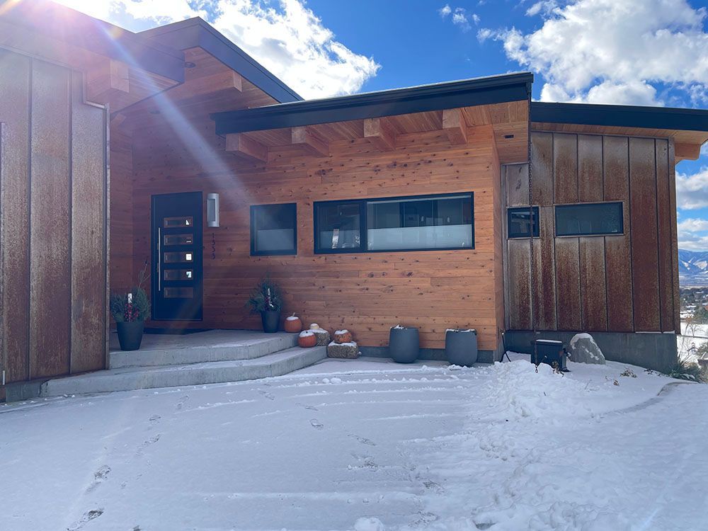 Modern wood-paneled home with a black door, snowy ground, and clear blue sky.