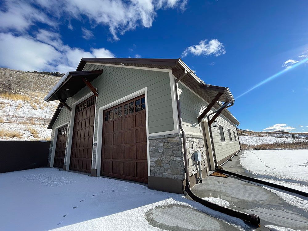 A green, single-story garage with brown carriage-style doors and stone accents, set against a snowy landscape.