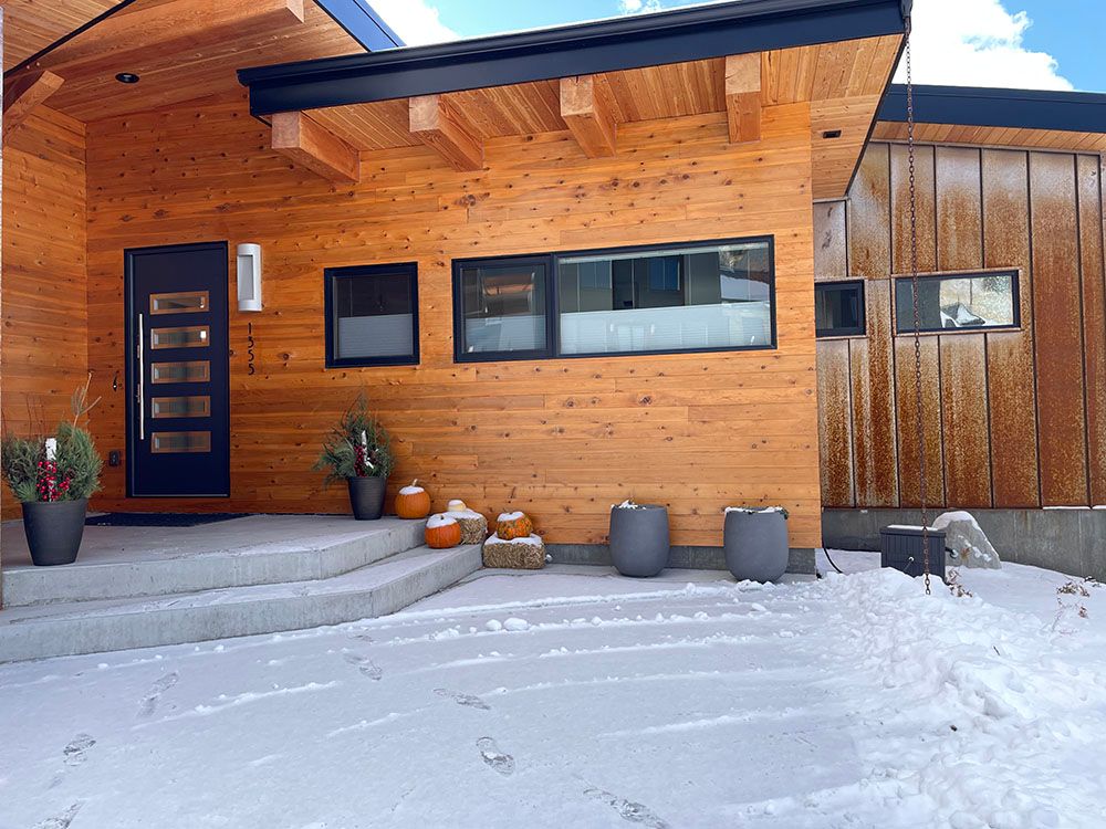 A modern home entrance with warm wood siding, a dark front door, and gray stone steps, surrounded by snow.