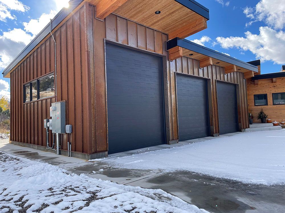 A modern wooden garage with three dark gray roll-up doors, set against a snowy landscape under a blue sky.