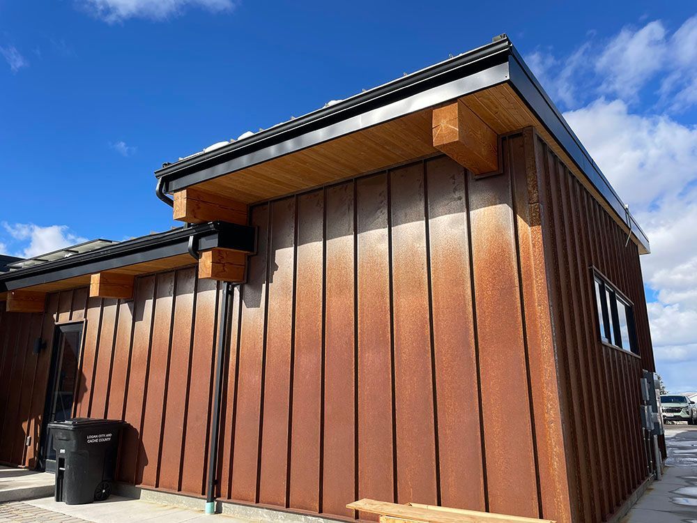 A building exterior with vertical rusted metal siding, wooden support beams, and a dark roof under a blue sky.