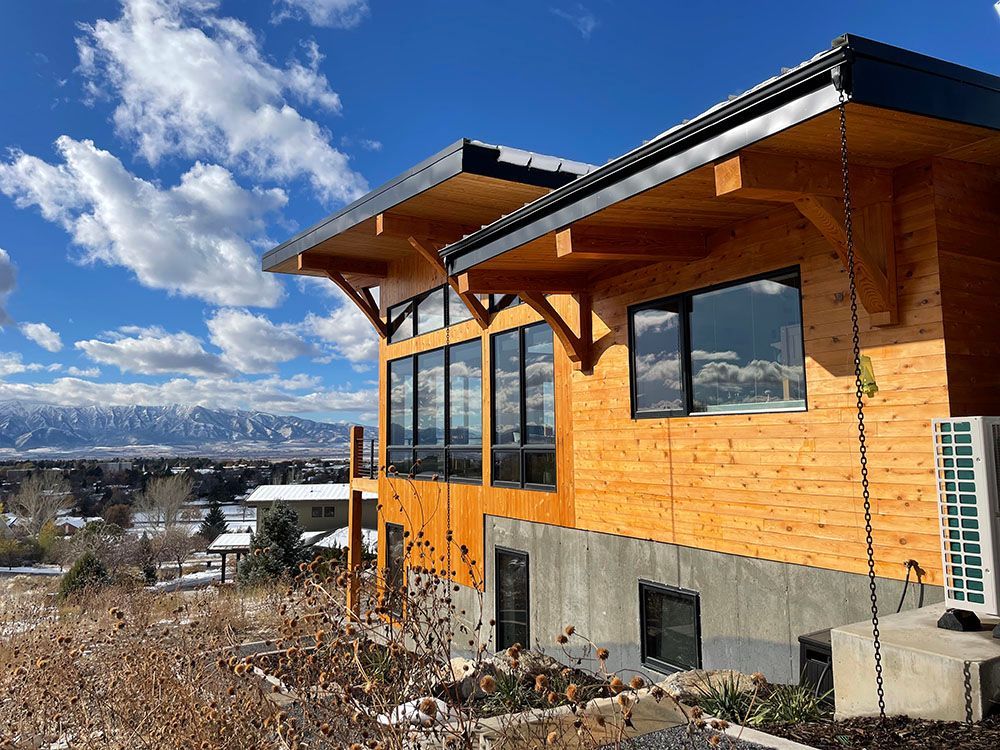 A modern wooden house with large windows sits on a hillside overlooking a snowy mountain landscape under a blue sky.