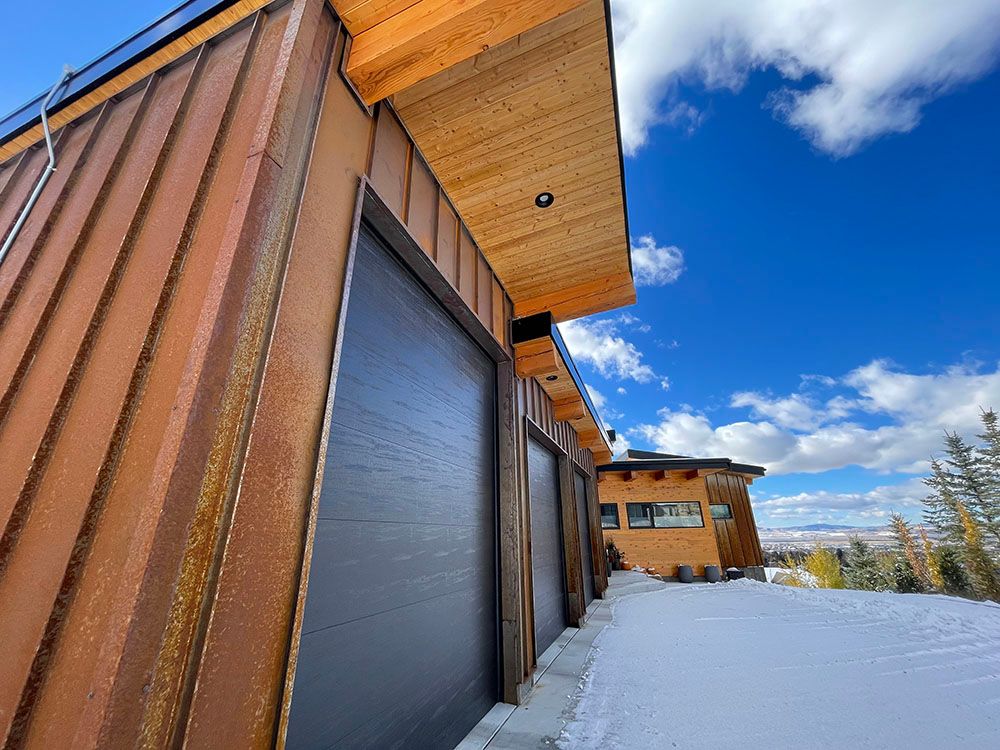 A modern house with rust-colored siding and a wooden overhang sits beside snowy ground under a bright blue sky.