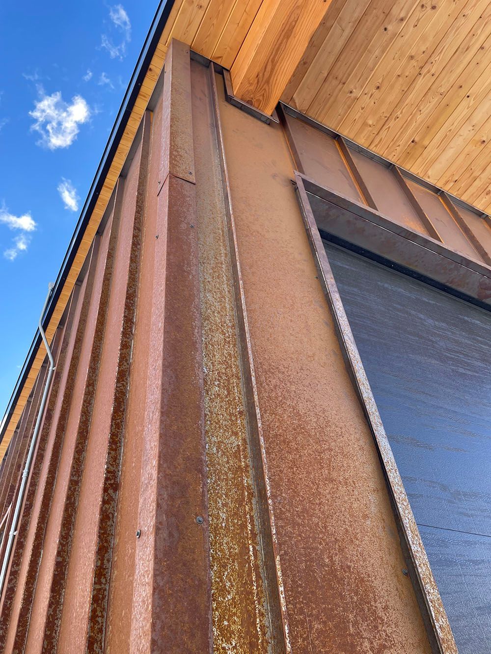 A low-angle view of a building corner with rusted, vertical metal panel cladding and a wooden ceiling soffit above.