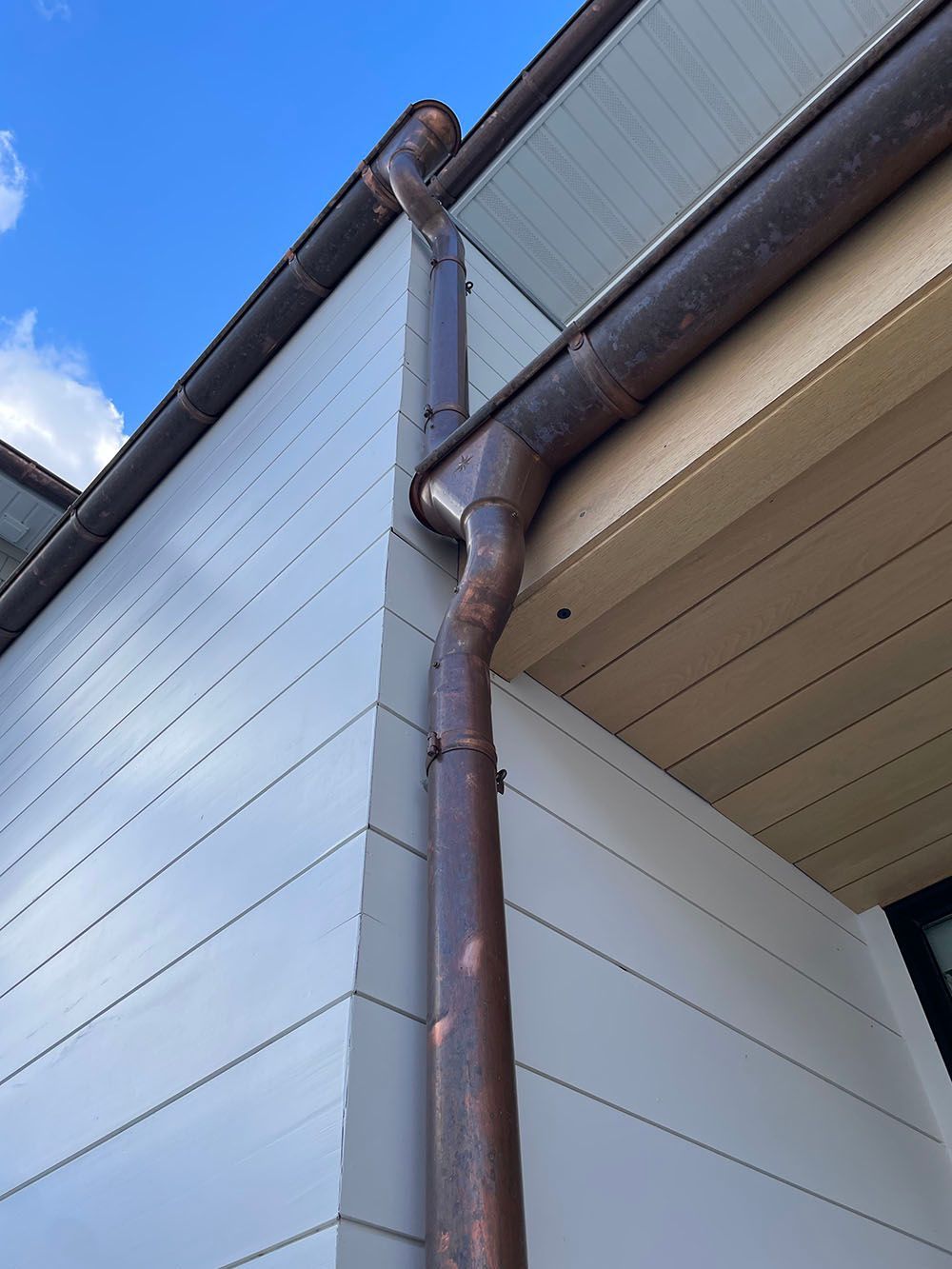 A rusted copper downspout attached to the corner of a building with light gray horizontal siding and a wood-paneled soffit.