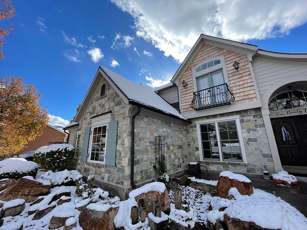 A stone house with a snowy roof, front yard, and light wood siding under a bright blue sky with scattered clouds.