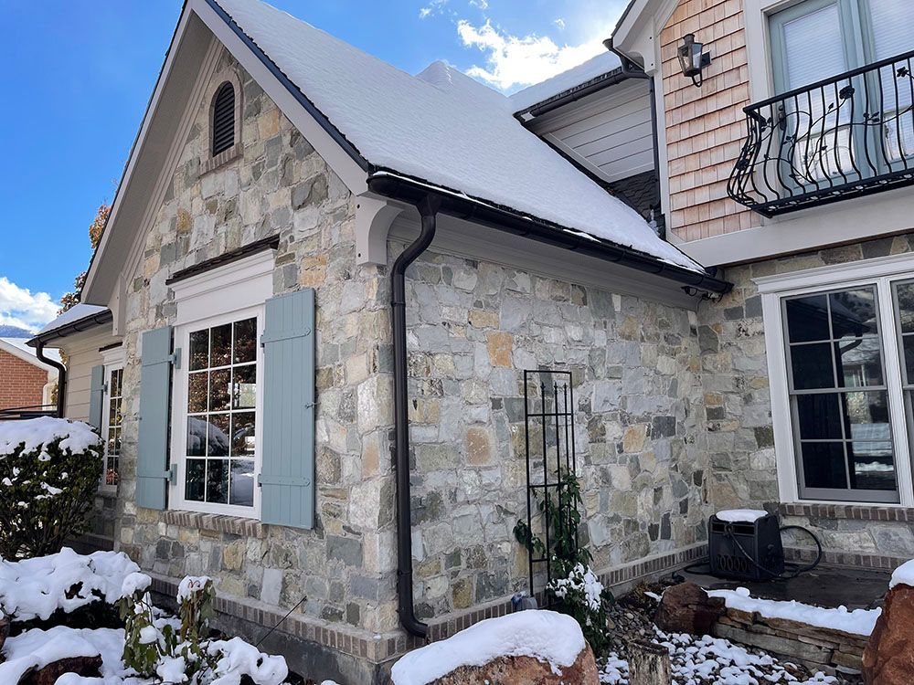 Exterior view of a stone house with light blue shutters and a snow-covered roof on a sunny day.
