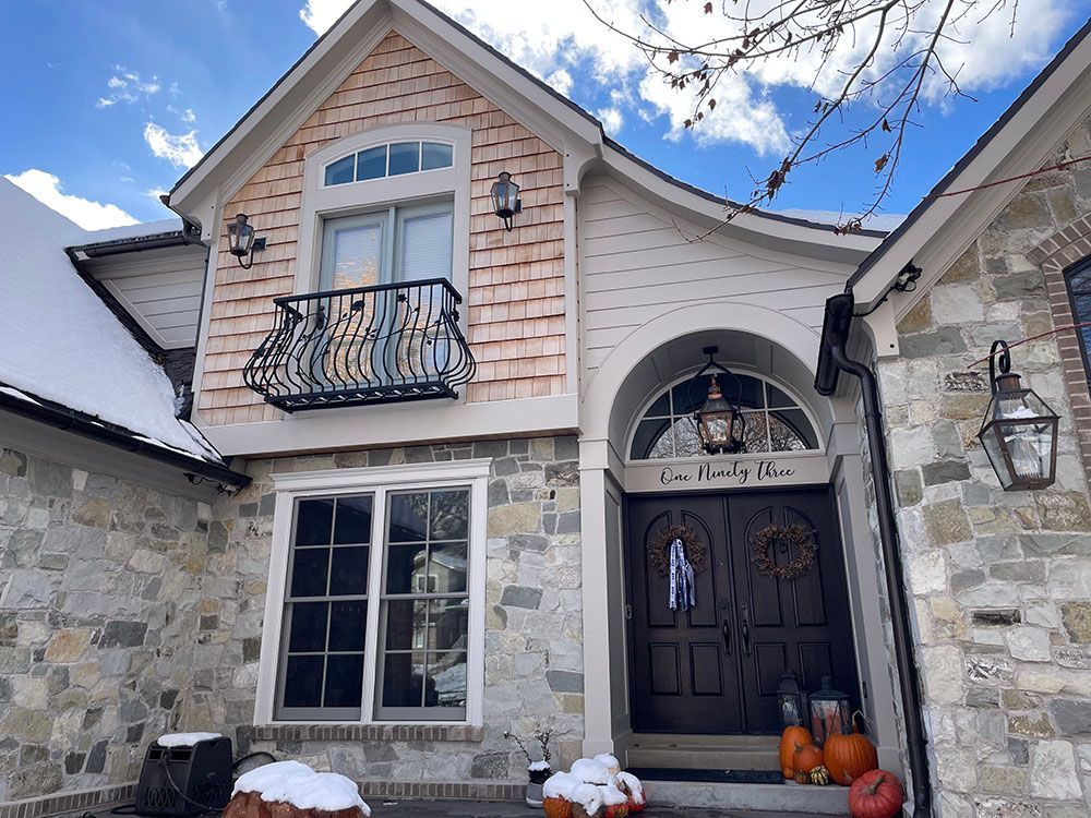 A two-story home exterior featuring light stone walls, tan cedar shakes on the upper level, a balcony, and front pumpkins.