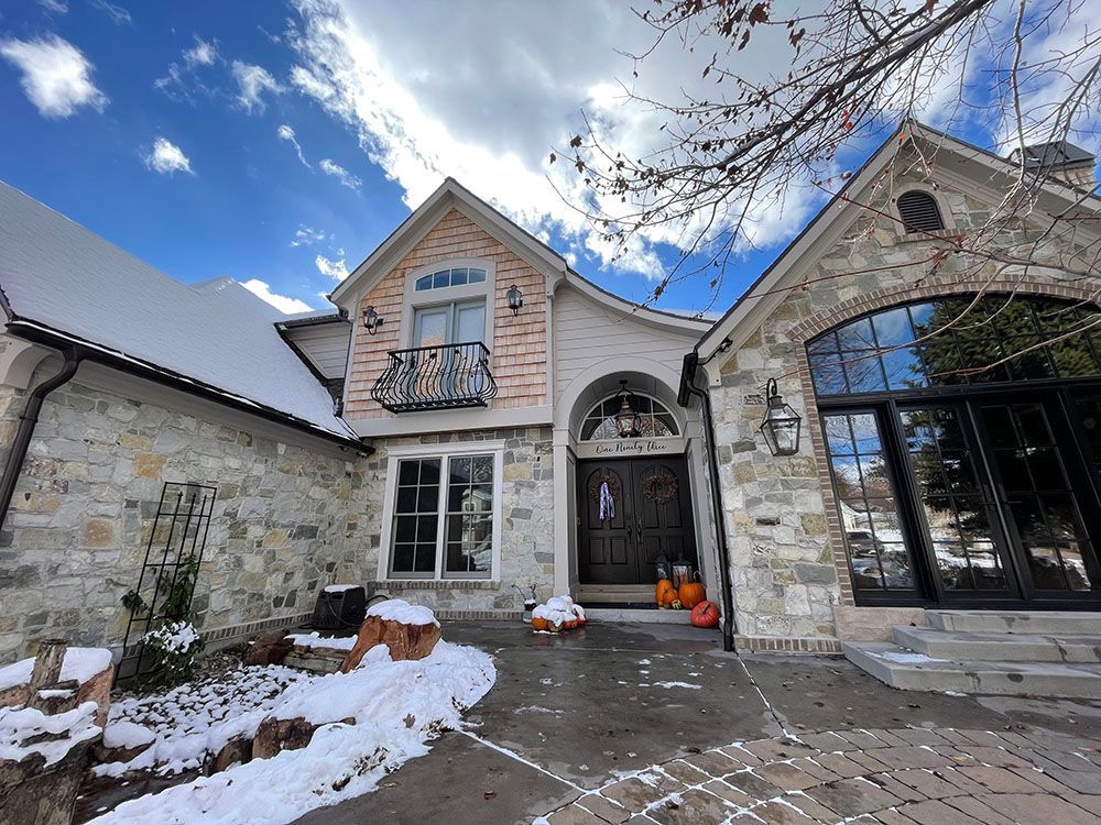 A stone-faced house with a dark front door, balcony, and snowy ground under a bright blue sky.