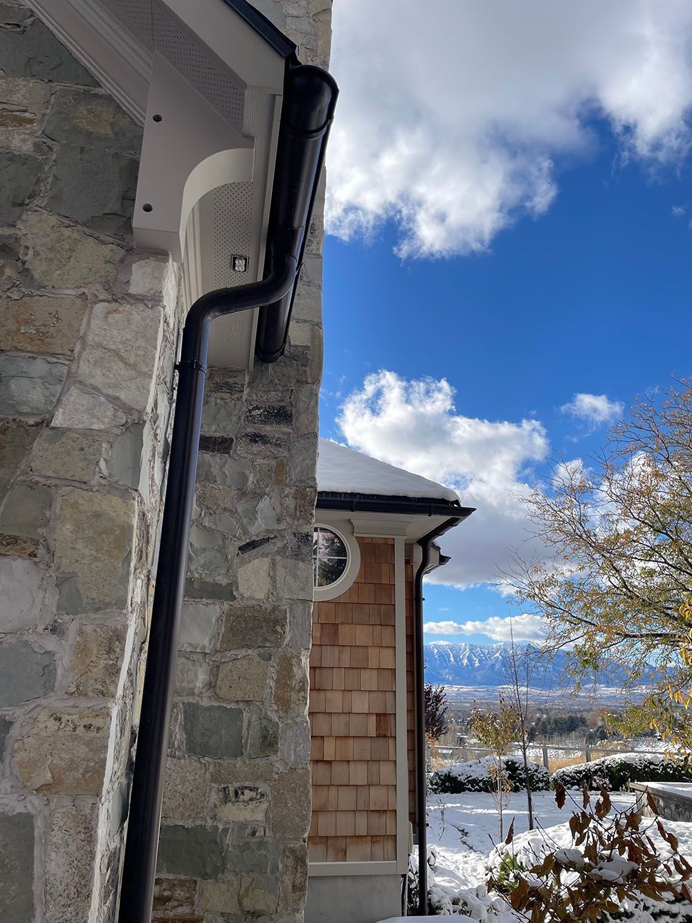 A close-up of a black gutter and downspout on a stone wall, with a view of a snowy landscape and mountains beyond.