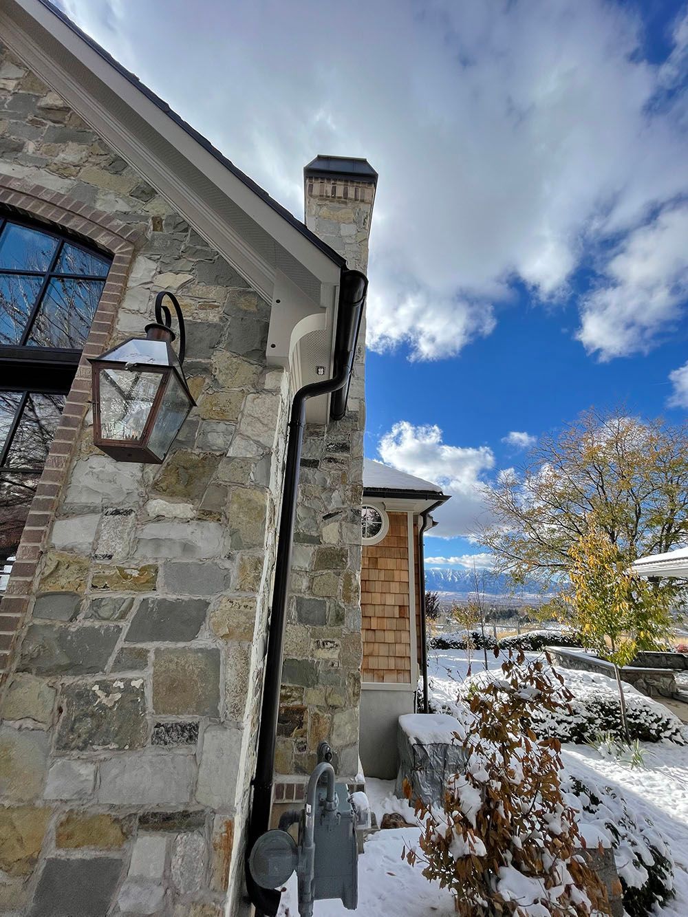 A stone exterior wall of a house with a dark lantern, a gutter, and a chimney, set against a snowy landscape and blue sky.