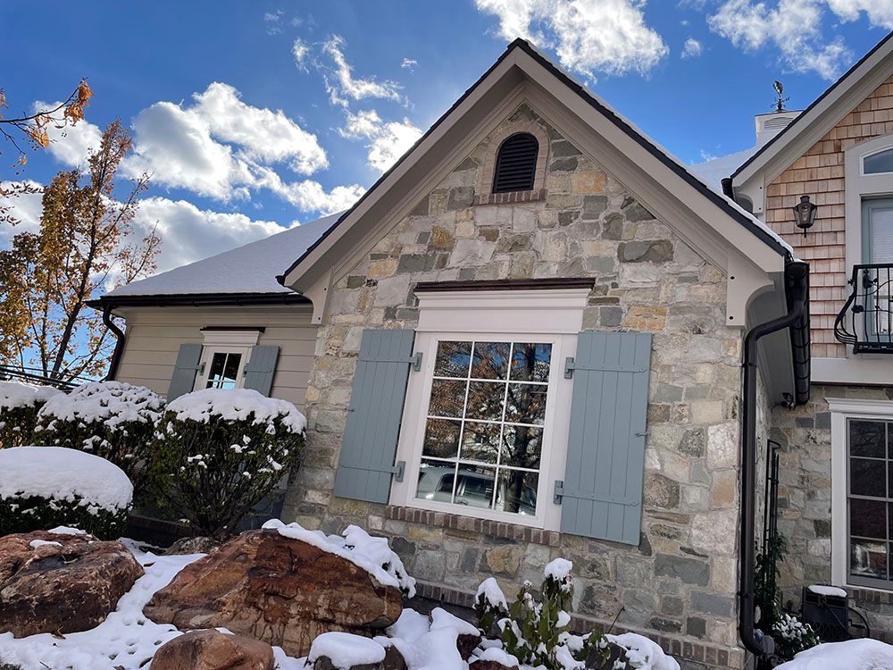 A stone-walled house exterior with blue shutters on a multi-pane window, featuring a snow-covered yard and bright sky.