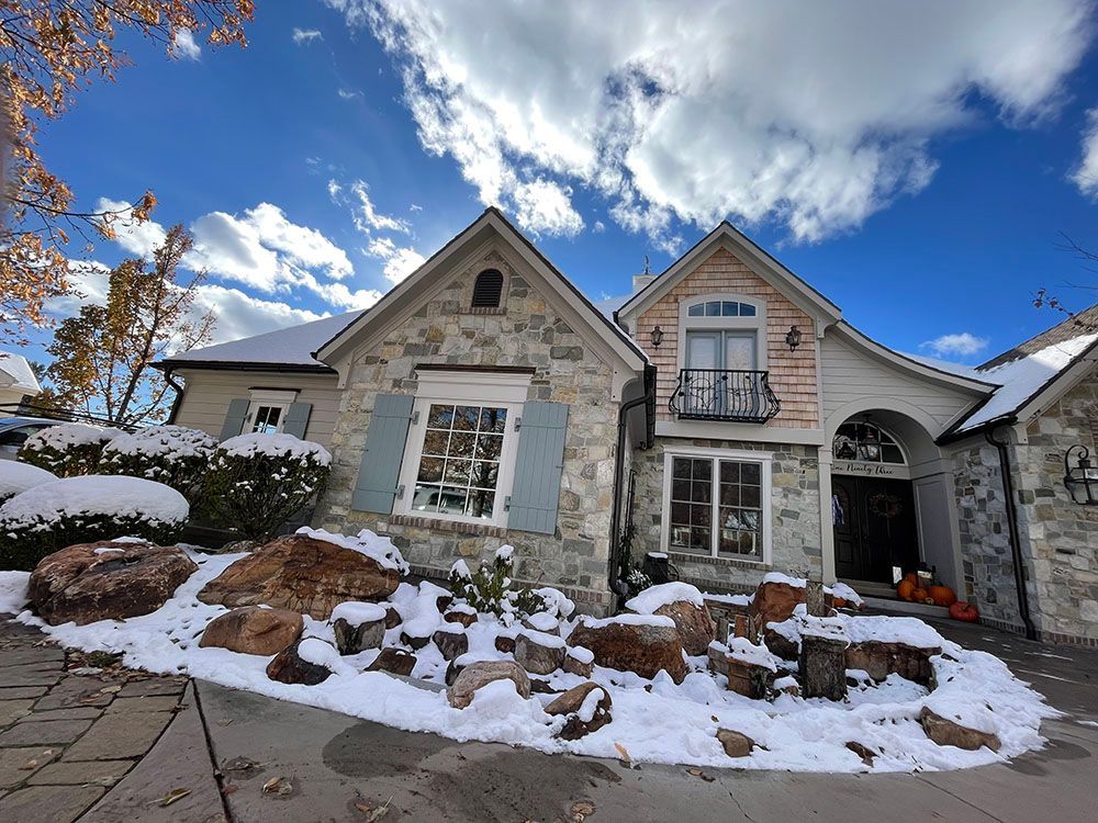 A two-story stone house with snow-covered rocks in the front garden under a bright blue sky.