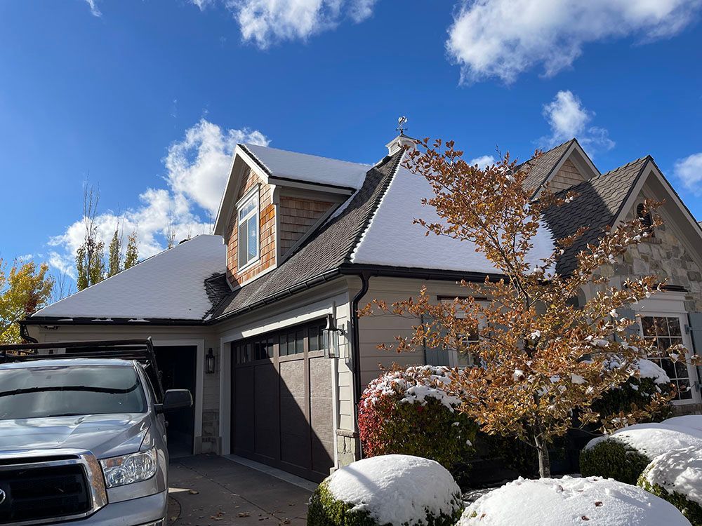 A silver truck is parked in the driveway of a suburban home featuring a roof partially covered in fresh snow.