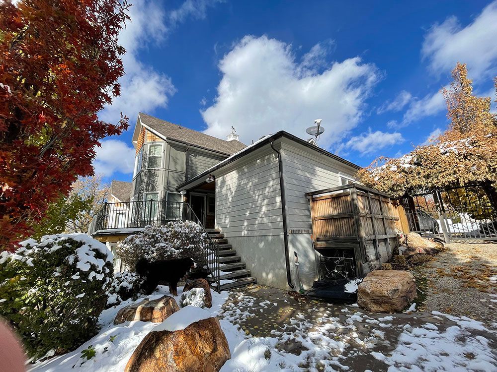 A white house with a multi-level roof and stairs, partially covered in snow under a bright blue sky with fall foliage.