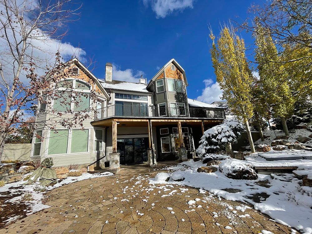 A large, multi-story house with grey siding and wooden accents stands under a blue sky, surrounded by light snow.