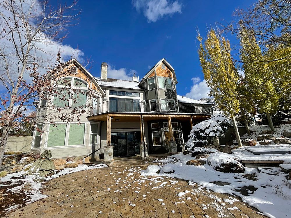 A multi-story house with stone and wood siding sits in a snowy yard under a bright blue sky with scattered clouds.