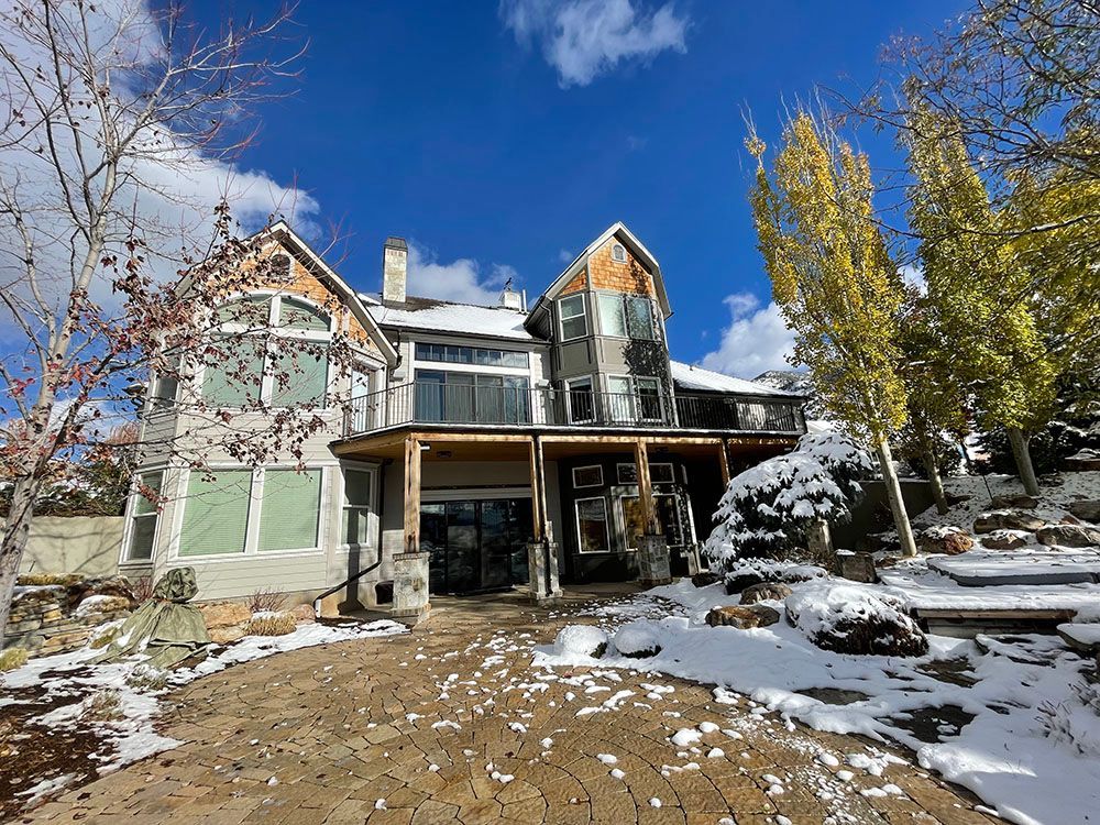 A multi-story house with stone and wood siding sits under a bright blue sky, surrounded by snow-covered ground and trees.