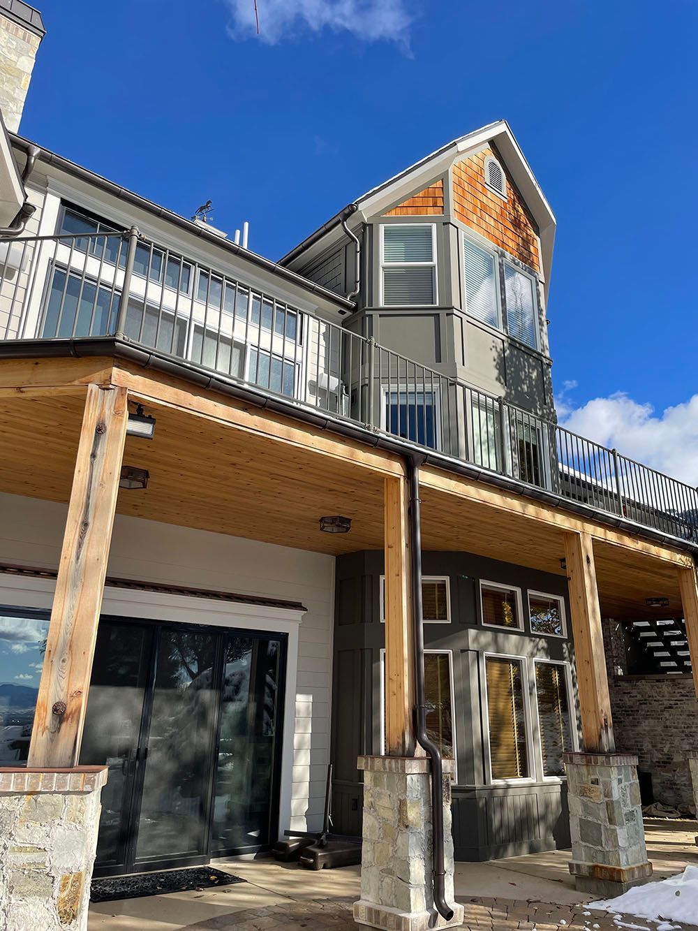 A three-story home with a covered wooden porch, grey siding, stone pillars, and a balcony against a blue sky.