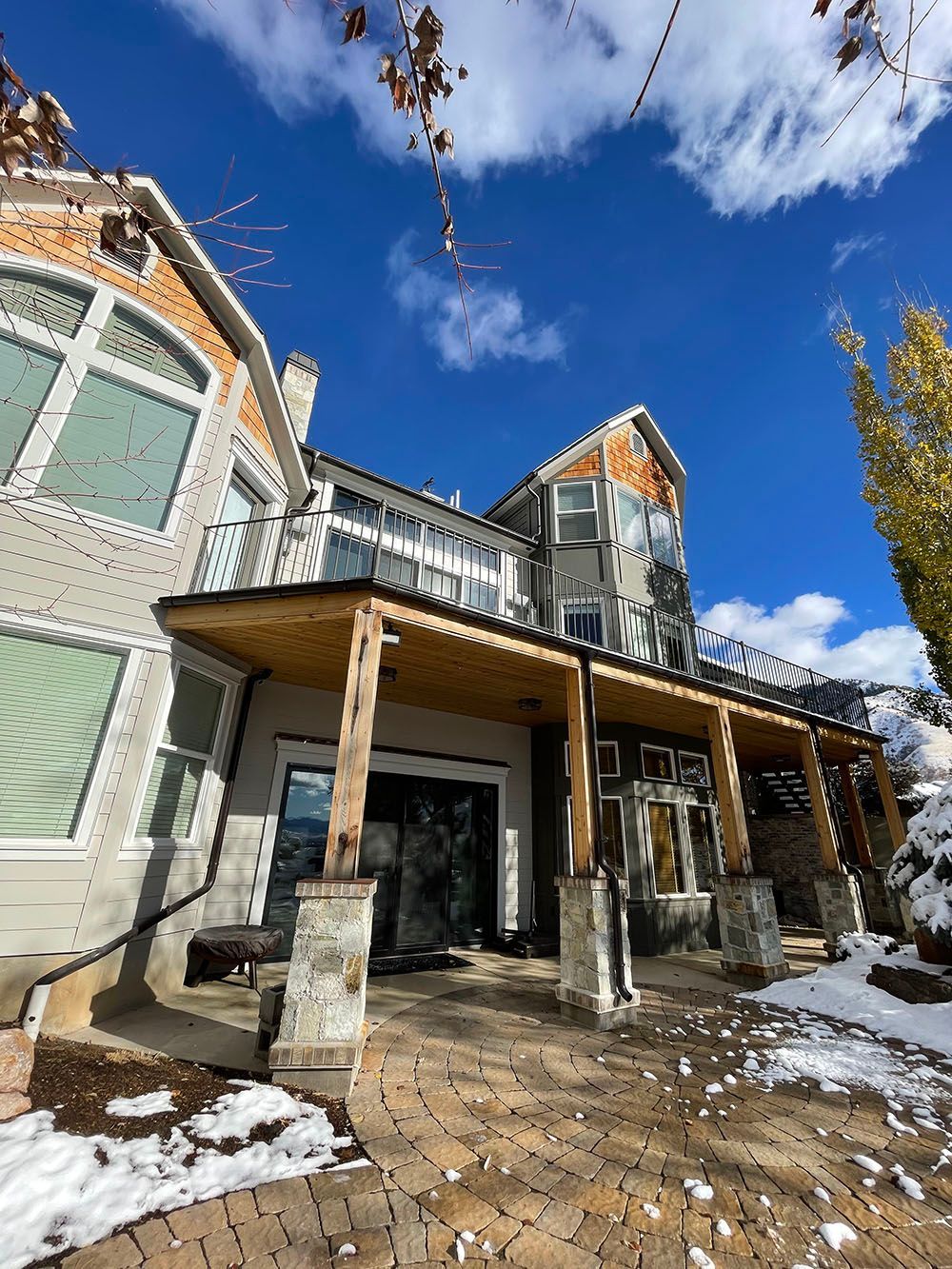 A low-angle view of a multi-story house with a wooden patio, stone columns, and snow-covered ground under a blue sky.