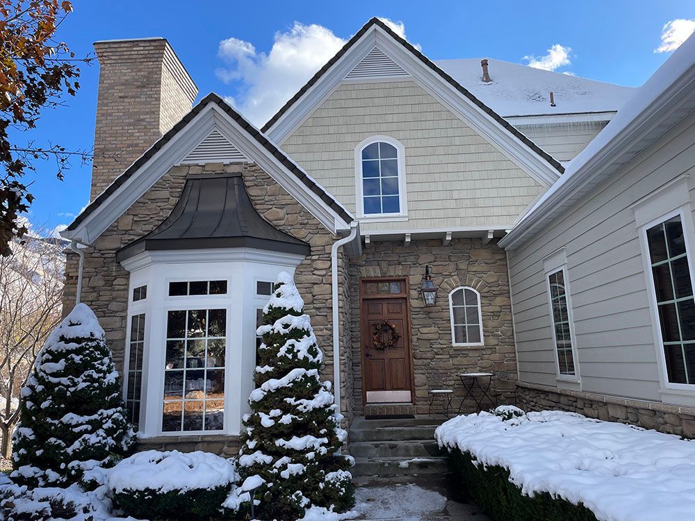 A stone and siding house in winter with snowy trees and a wooden front door under a clear blue sky.