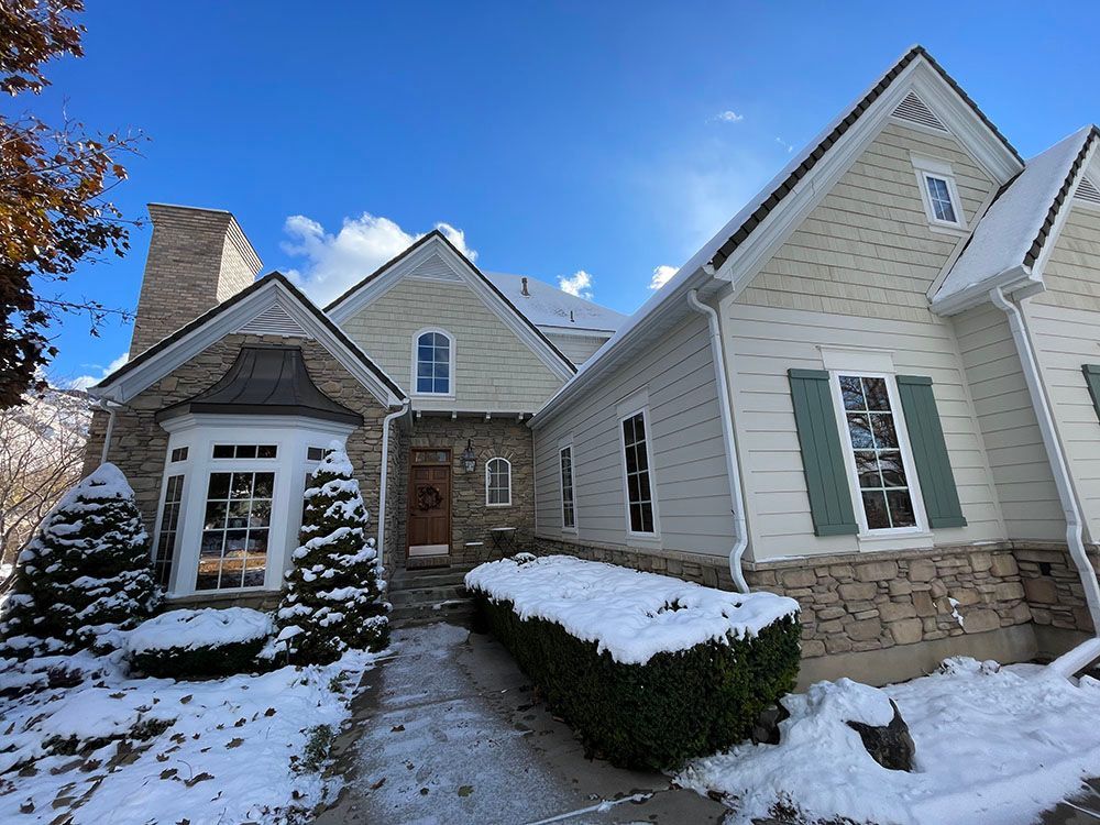 A winter scene of a light-colored home with stone accents, green shutters, and a snow-covered front yard and path.