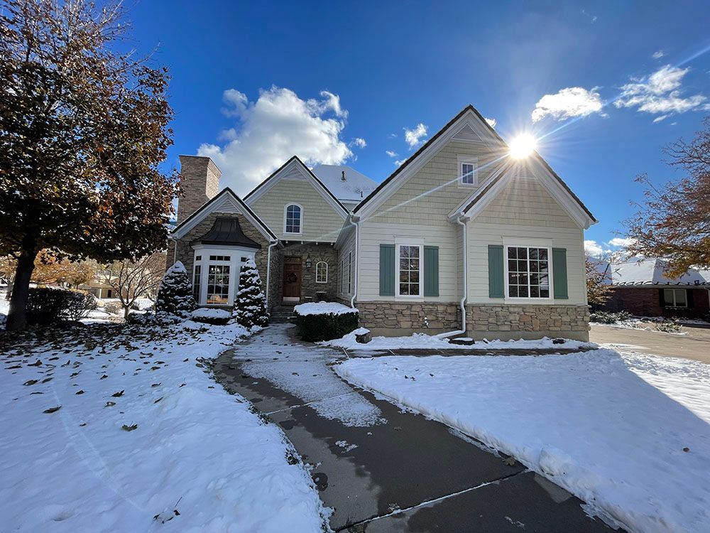A light-colored house with stone accents, a snow-covered yard, and a clear blue sky on a sunny winter day.