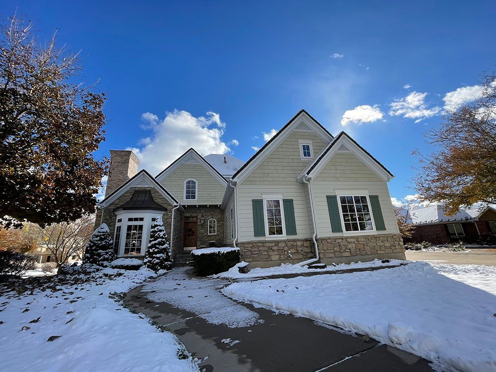 A two-story light green home with stone siding, dark shutters, and a snow-covered front yard under a bright blue sky.
