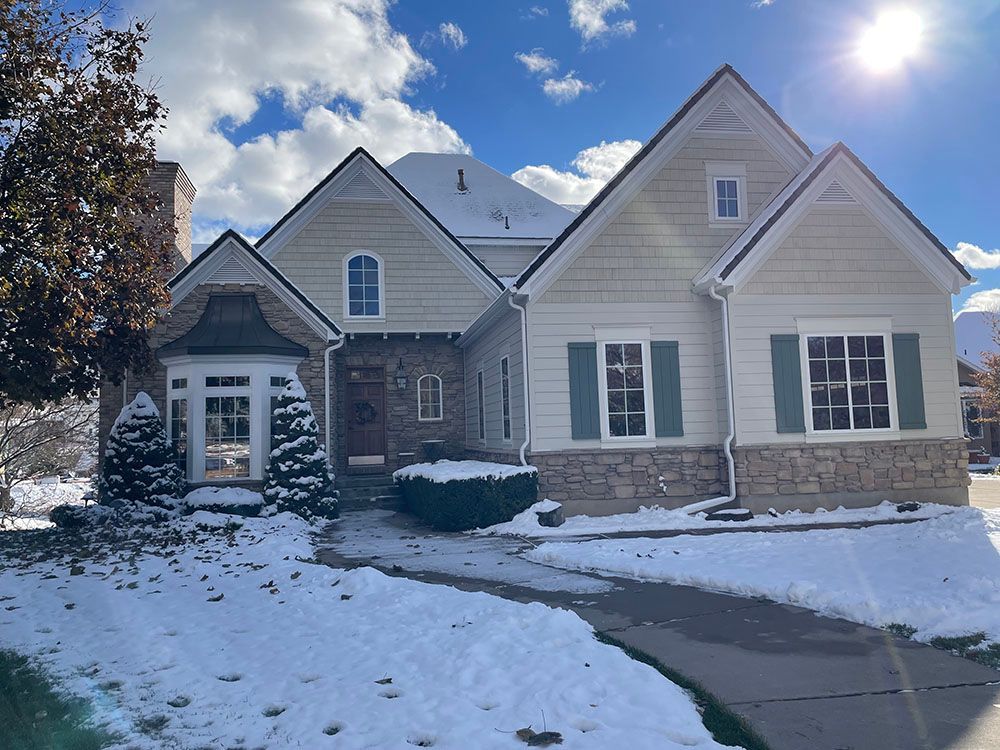 A two-story suburban home with light siding, a stone facade, and green shutters, partially covered in a dusting of snow.