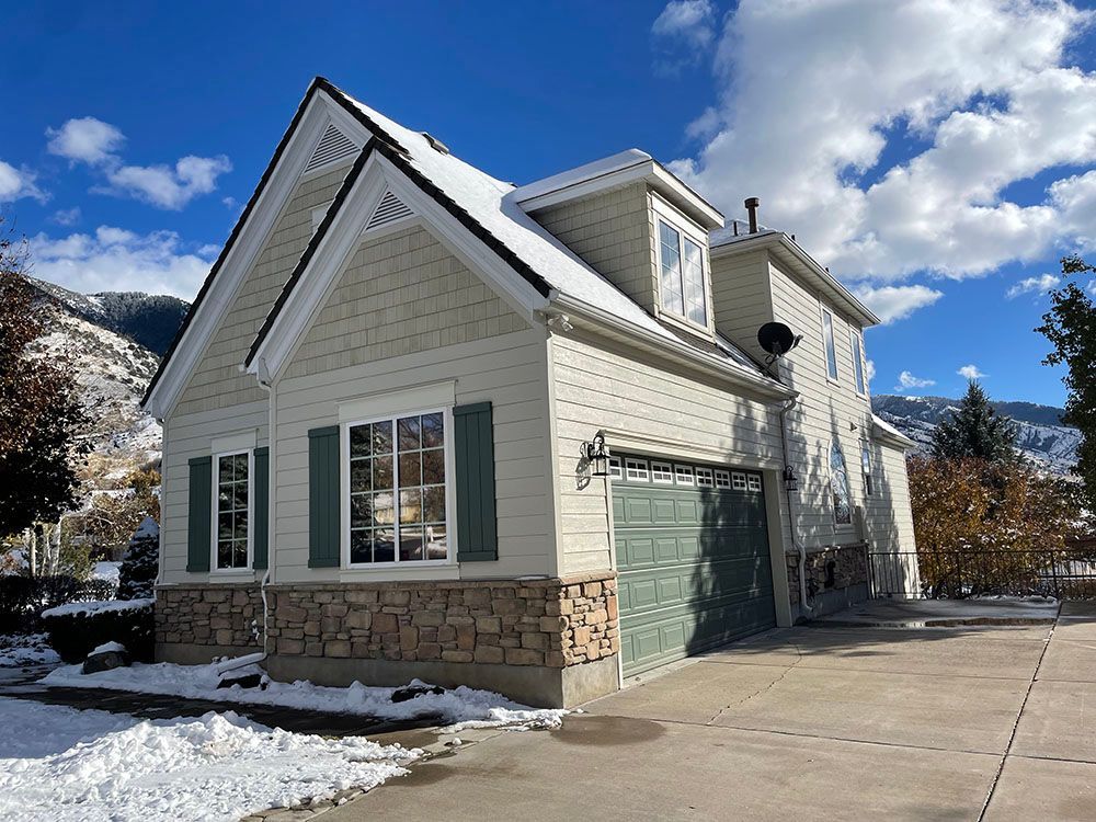 A two-story house with light beige siding, stone accents, and a green garage door, set against a snowy mountain backdrop.