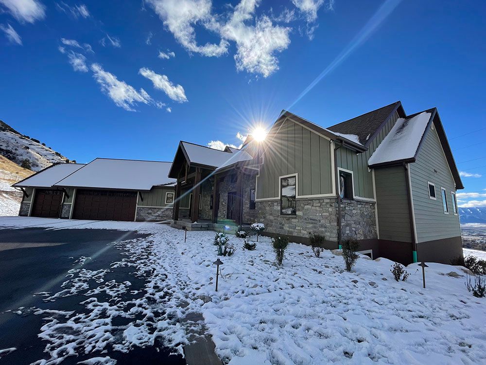 A modern house with a mix of sage green siding and stone veneer, surrounded by snow under a bright, sunny blue sky.