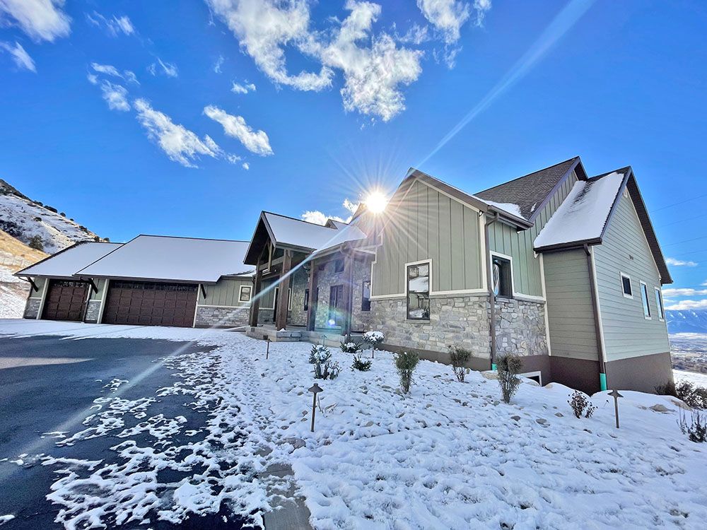 A modern house with gray siding and stone accents under a sunny blue sky, surrounded by a light covering of snow.