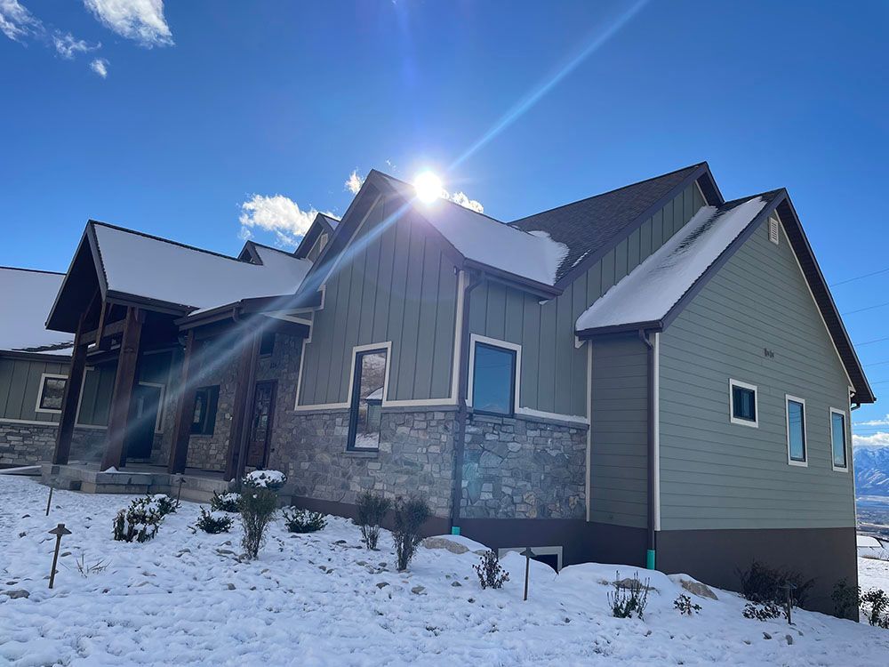 A modern two-story home with gray siding and stone accents, partially covered in snow, set against a bright blue sky.
