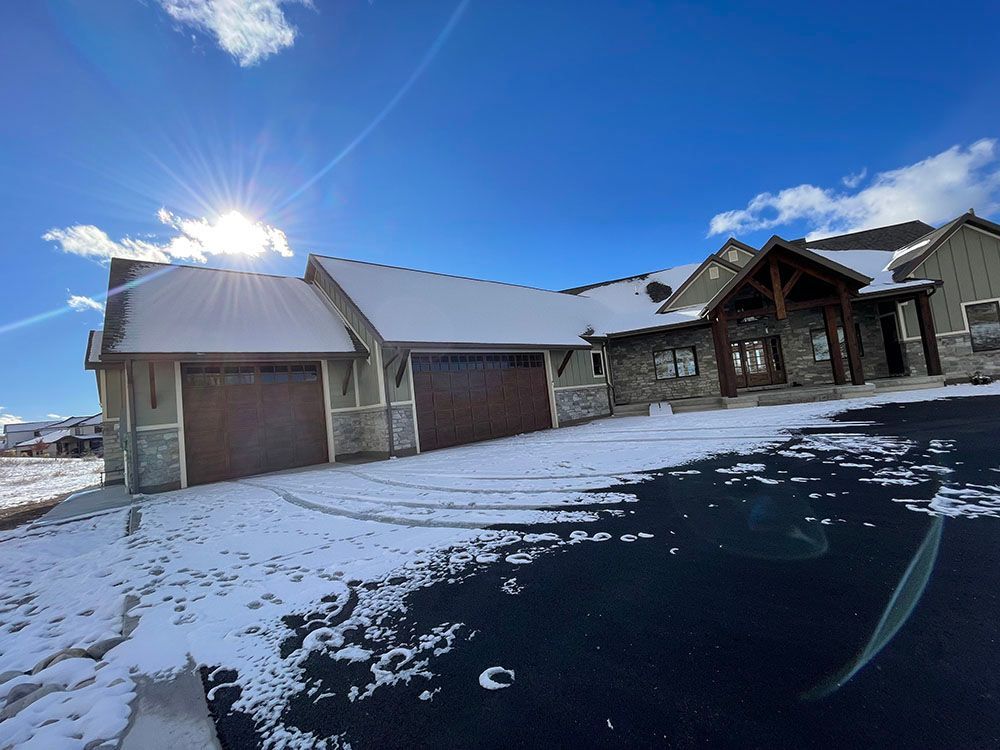 A residential house with a multi-car garage, stone and siding exterior, and snow-covered roof under a bright, sunny sky.