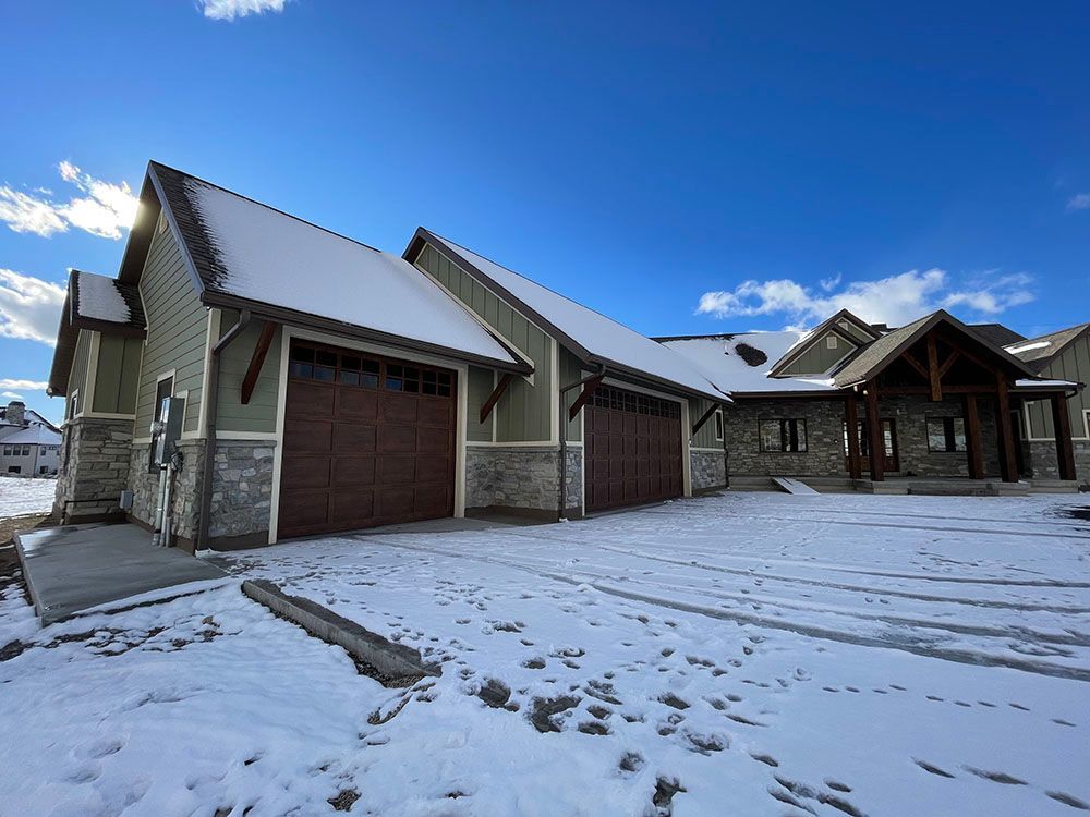 A modern home with a two-car garage, stone and green siding, and snowy ground under a bright blue sky.