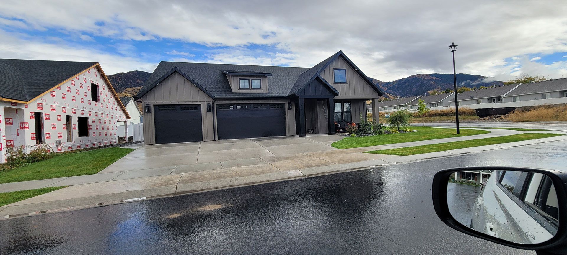 A modern grey suburban home with dark garage doors sits next to a house under construction with visible insulation.